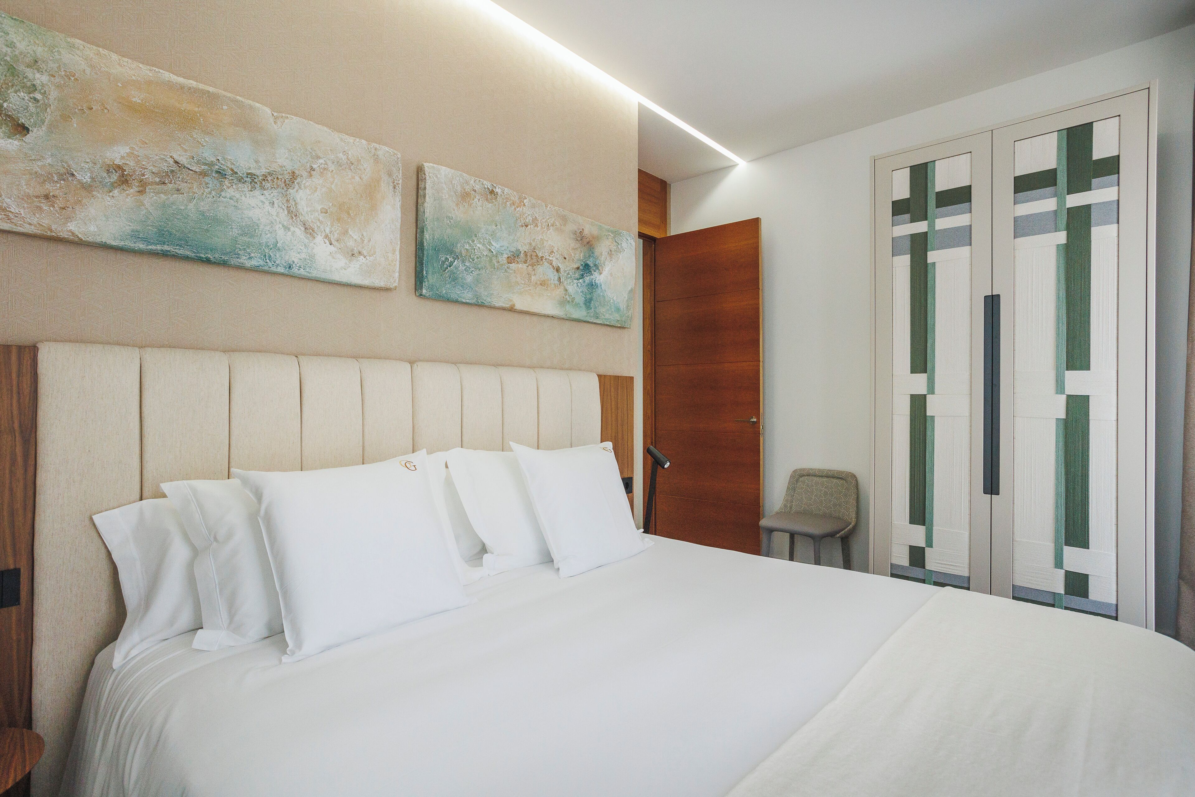 A neatly made bed with white linens and a headboard, a wooden door, and two abstract wall paintings.