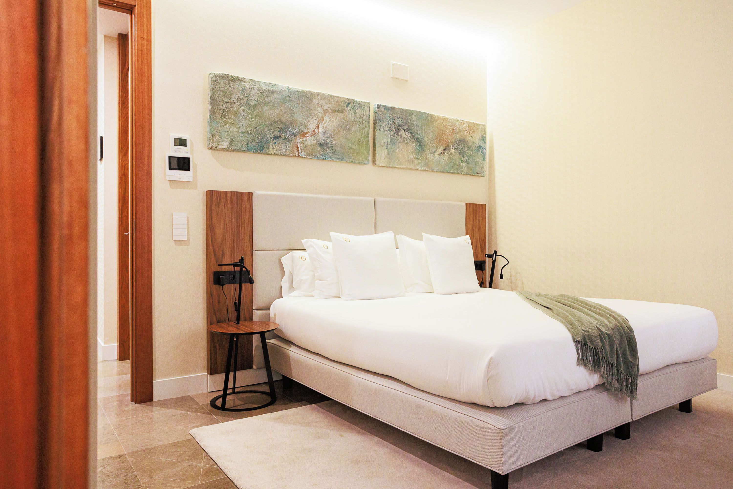Habitación de hotel con cama, mesitas de noche, termostato fijado a la pared y arte abstracto en la pared.
