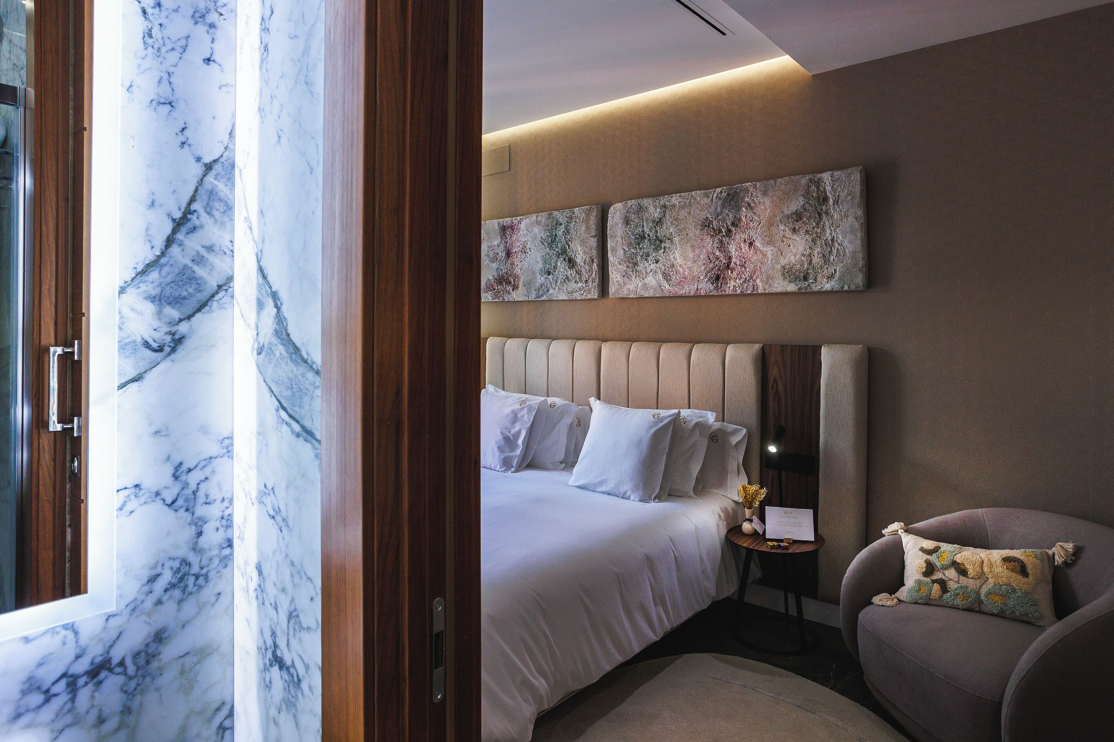 A modern hotel room with a marble wall, a large bed with white bedding, a headboard, a side table with a lamp, and a plush armchair.