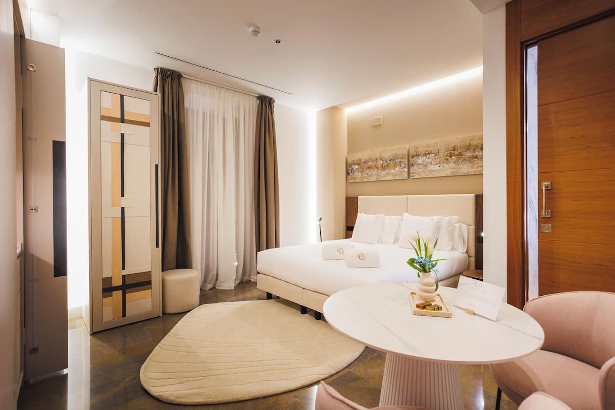 Una habitación de hotel moderna con una cama grande, una mesa redonda, un rincón de descanso y una puerta con panel de vidrio.