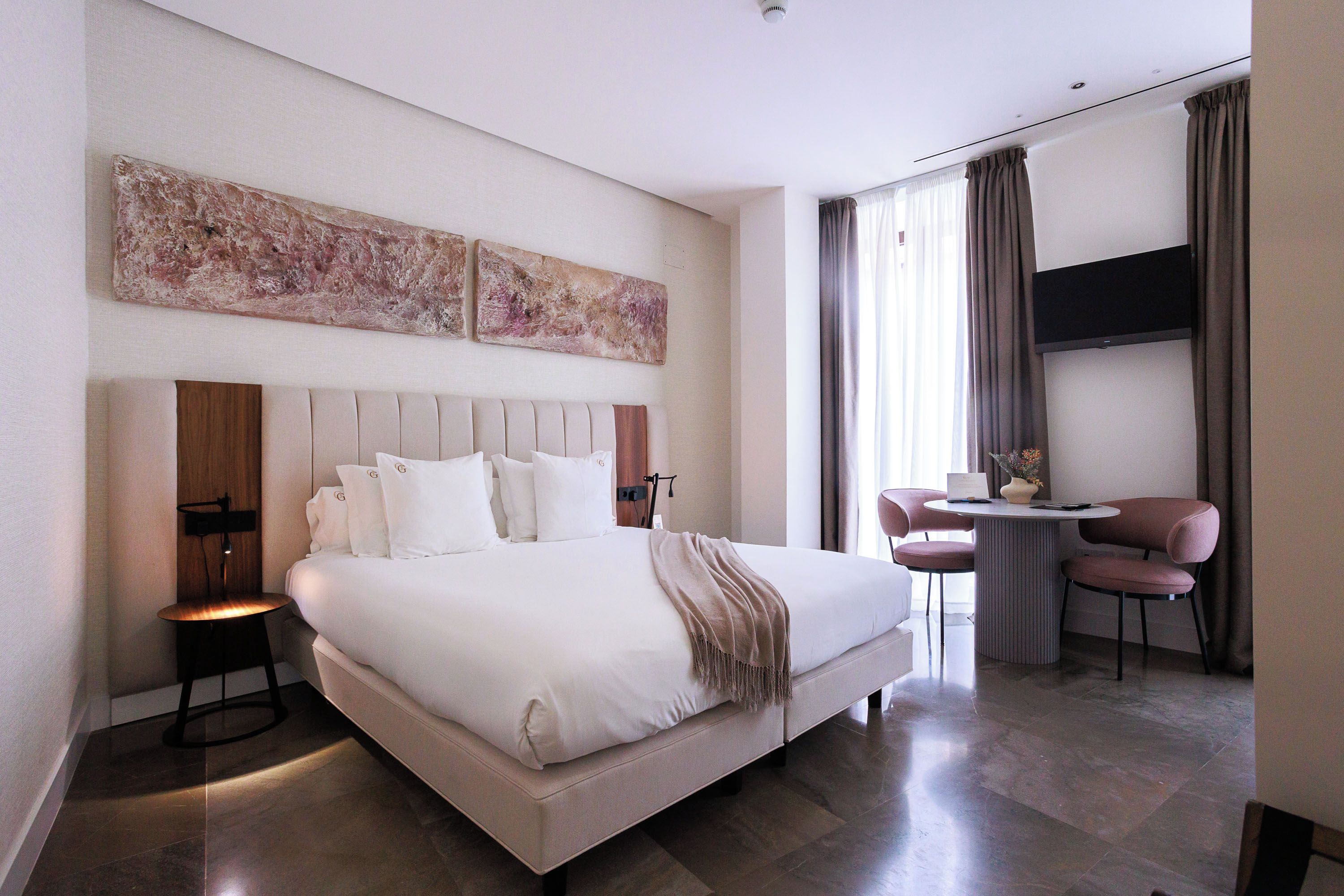 A modern hotel room with a large bed, a bedside table, a small round table with chairs, and a flat-screen TV mounted on the wall.