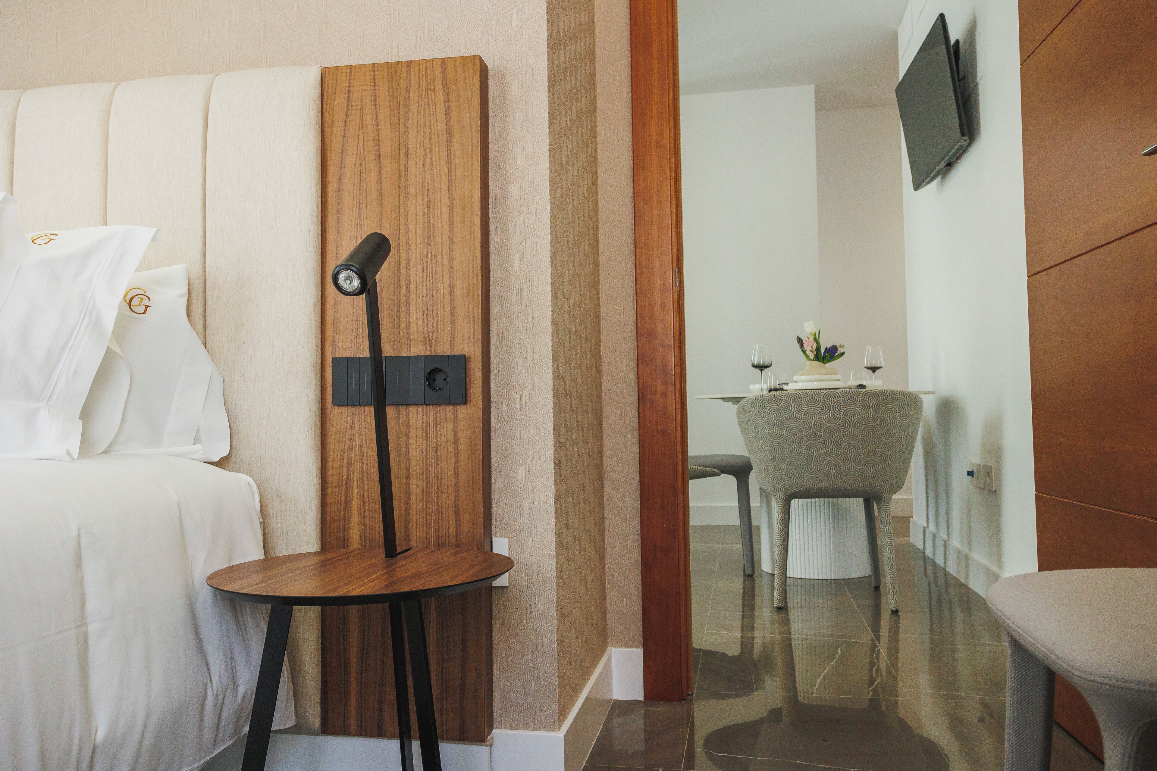 Una habitación de hotel con una cama, una mesita de madera, una silla, una mesa de comedor con sillas y un televisor de pantalla plana montado en la pared.
