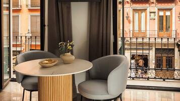 A modern interior with a round table, two chairs, a flat-screen TV, and a view of a red building with balconies.