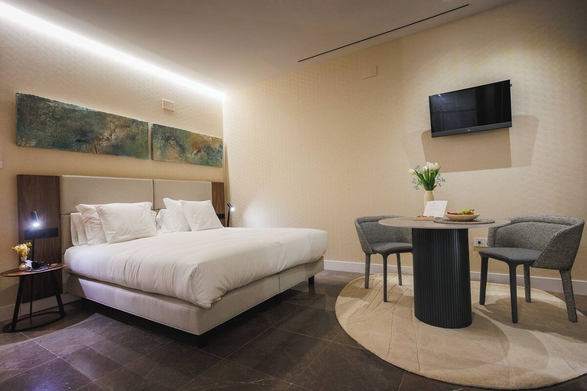 Una habitación de hotel moderna con una cama grande, una mesa de comedor redonda, dos sillas, un televisor de pantalla plana y arte abstracto en las paredes.
