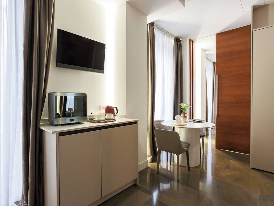 Habitación de hotel moderna con kitchenette, televisor de pantalla plana sobre la encimera, zona de comedor con mesa y sillas, y un amplio ventanal con cortinas.