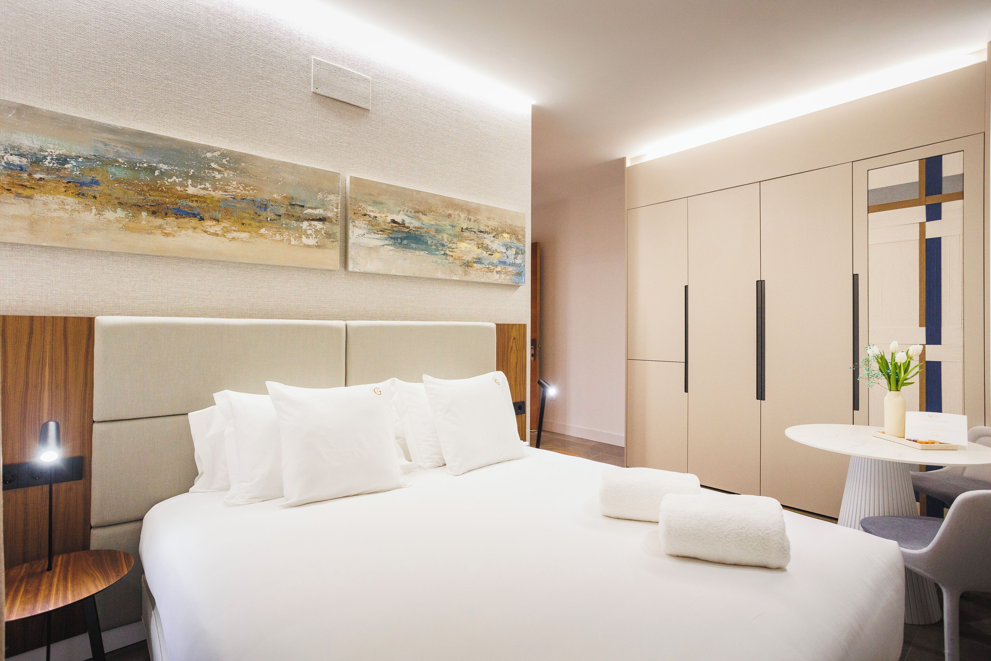A modern hotel room with a large bed, a bedside table, a small round table, and a wardrobe.