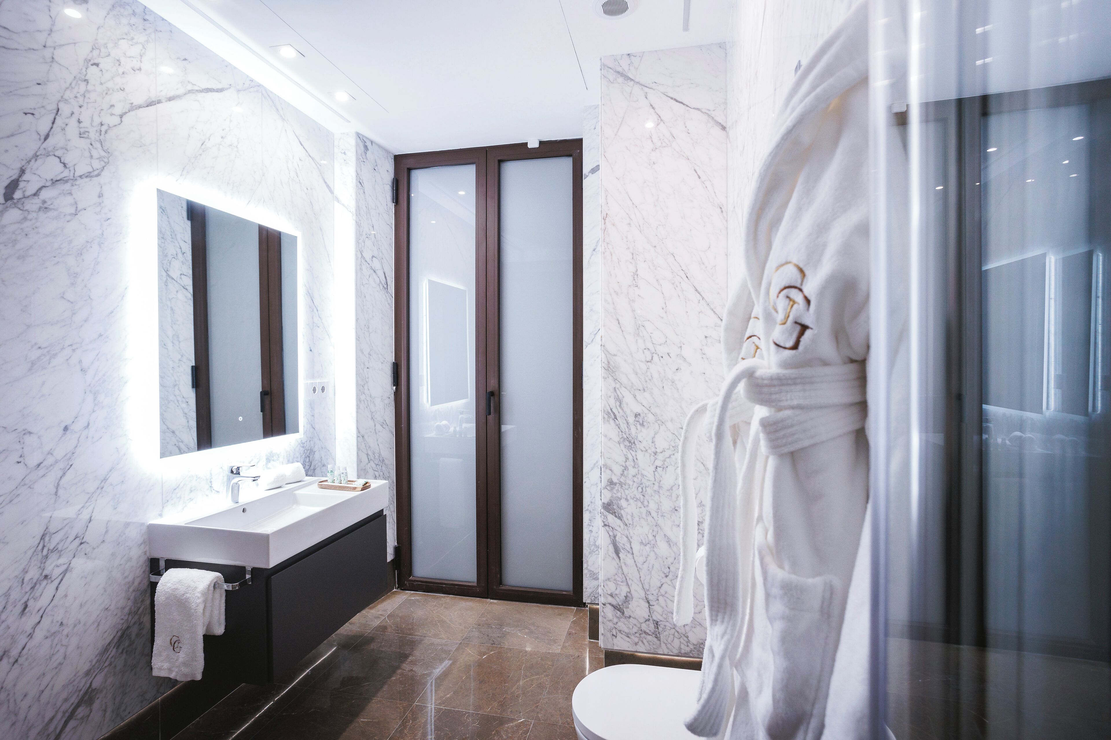 A modern bathroom with a marble wall, a large mirror, a white sink, and a glass shower enclosure.