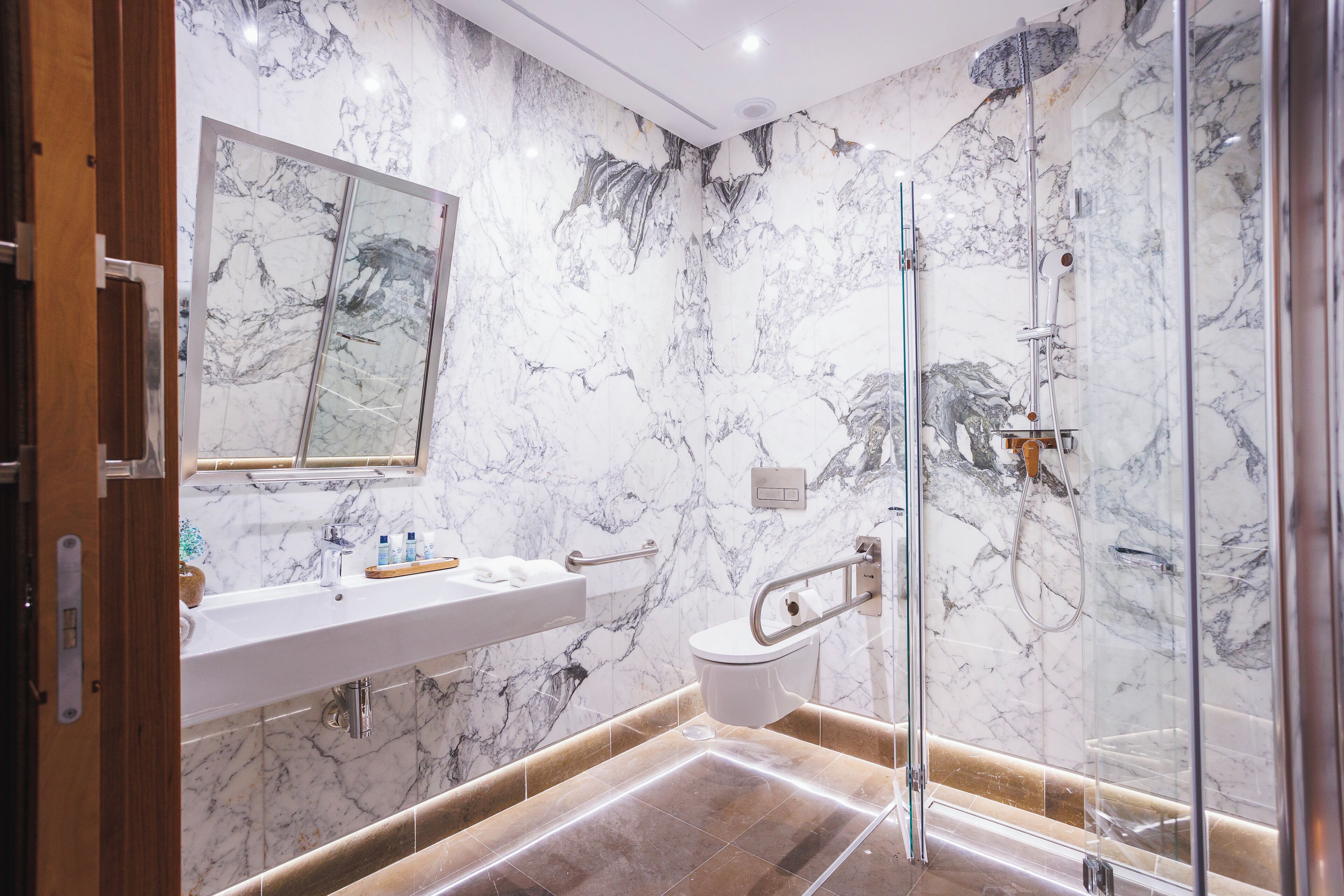 Un baño moderno con pared de mármol, cabina de ducha de vidrio, lavamanos blanco y puerta de madera.