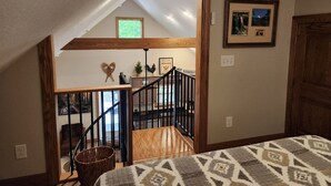A bedroom with a patterned bedspread, a wooden headboard, and a view of a staircase leading to a loft area with a window.