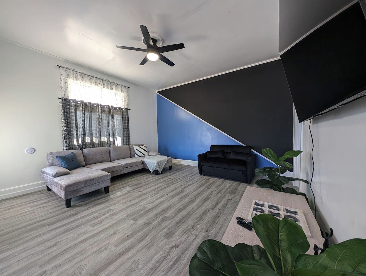 Un salon moderne avec un canapé d’angle gris, une petite table blanche, un canapé noir, un ventilateur de plafond et une grande plante.