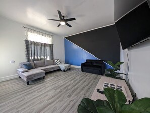 Una sala de estar moderna con un sofá seccional gris, una mesita blanca, un sofá negro, un ventilador de techo y una planta grande.