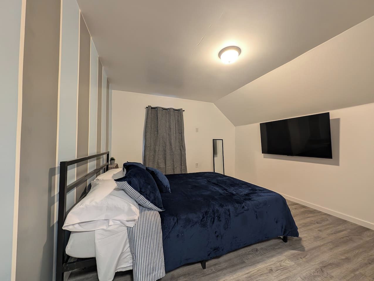 A bedroom with a bed, a bedside table, a flat-screen TV, and a window with curtains.