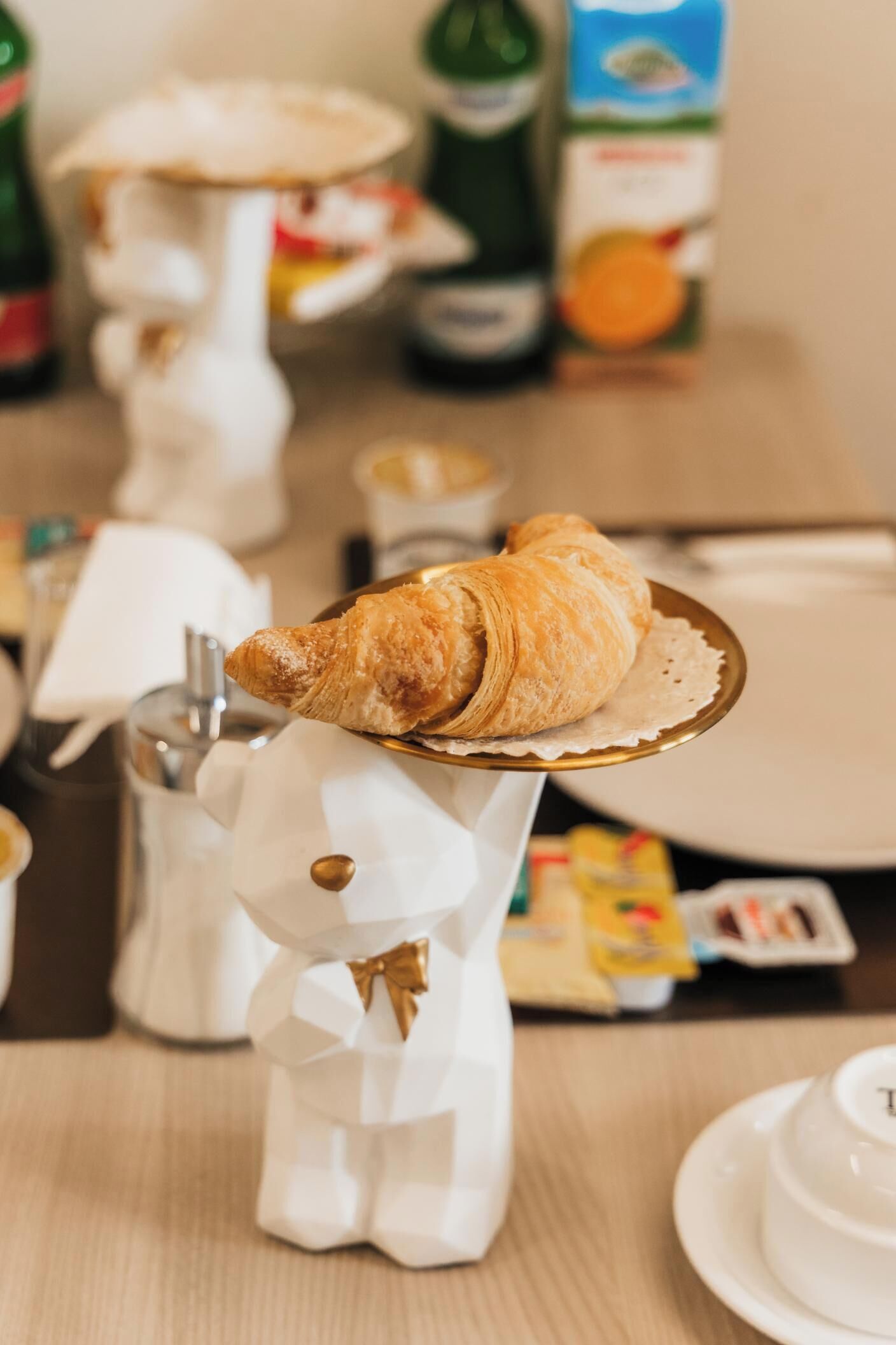 A breakfast setting with a croissant on a golden stand, a white ceramic cup, and a carton of orange juice.