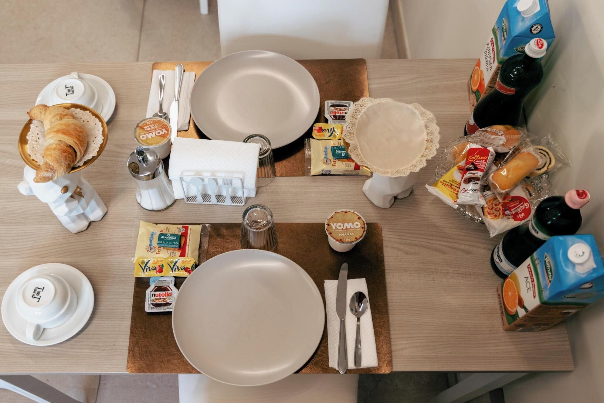 Een ontbijttafel met borden, bestek, een croissant en diverse etenswaren.