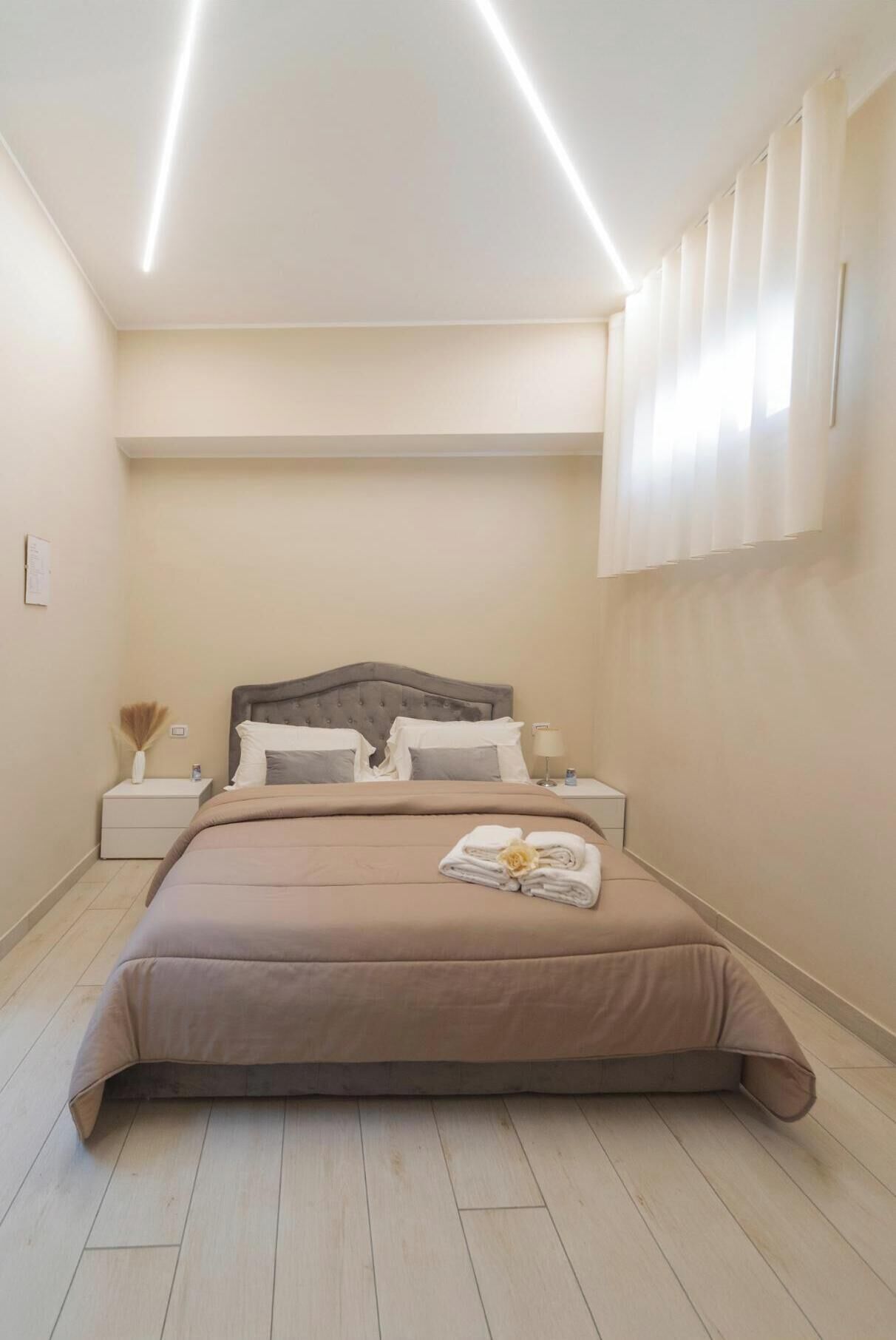 Un dormitorio con cama, mesitas de noche, luz empotrada en la pared y ventana con persianas.
