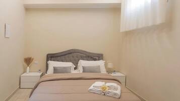 Un dormitorio con cama, mesitas de noche, luz empotrada en la pared y ventana con persianas.