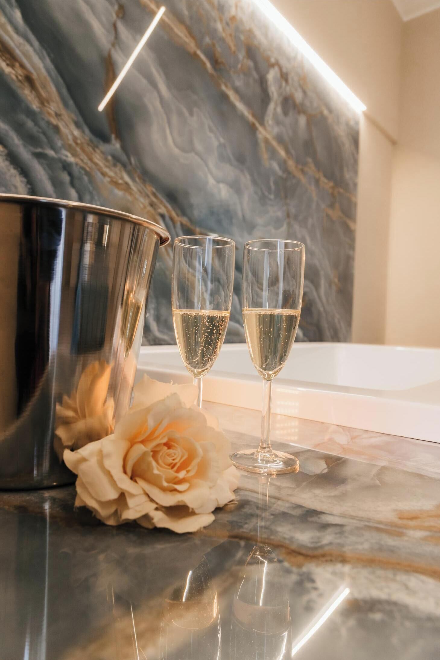 Une table surmontée d’un plateau de marbre, avec un seau à champagne et deux verres, une rose blanche et un luminaire moderne.