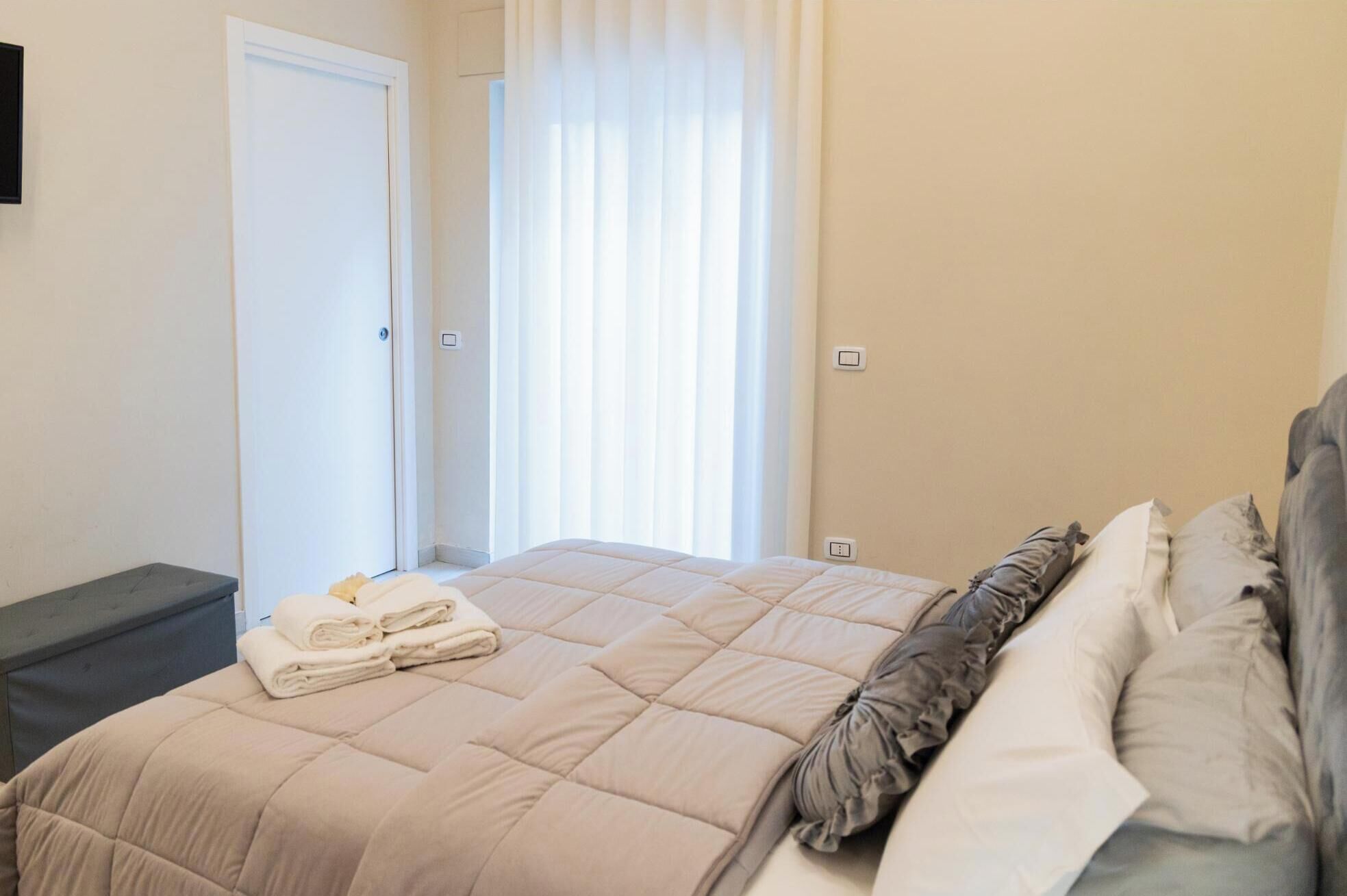 Uma cama bem arrumada com um edredom bege e travesseiros, uma mesinha de cabeceira com toalhas dobradas e uma janela com cortinas translúcidas permitindo a entrada de luz natural.