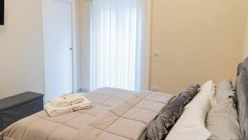 Una cama bien hecha con edredón y almohadas color beige, una mesita de noche con toallas dobladas y una ventana con cortinas traslúcidas que dejan entrar luz natural.