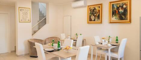 A dining area with a table set for four, white chairs, and a sofa. There are two paintings on the wall, a staircase, and an air conditioning unit.