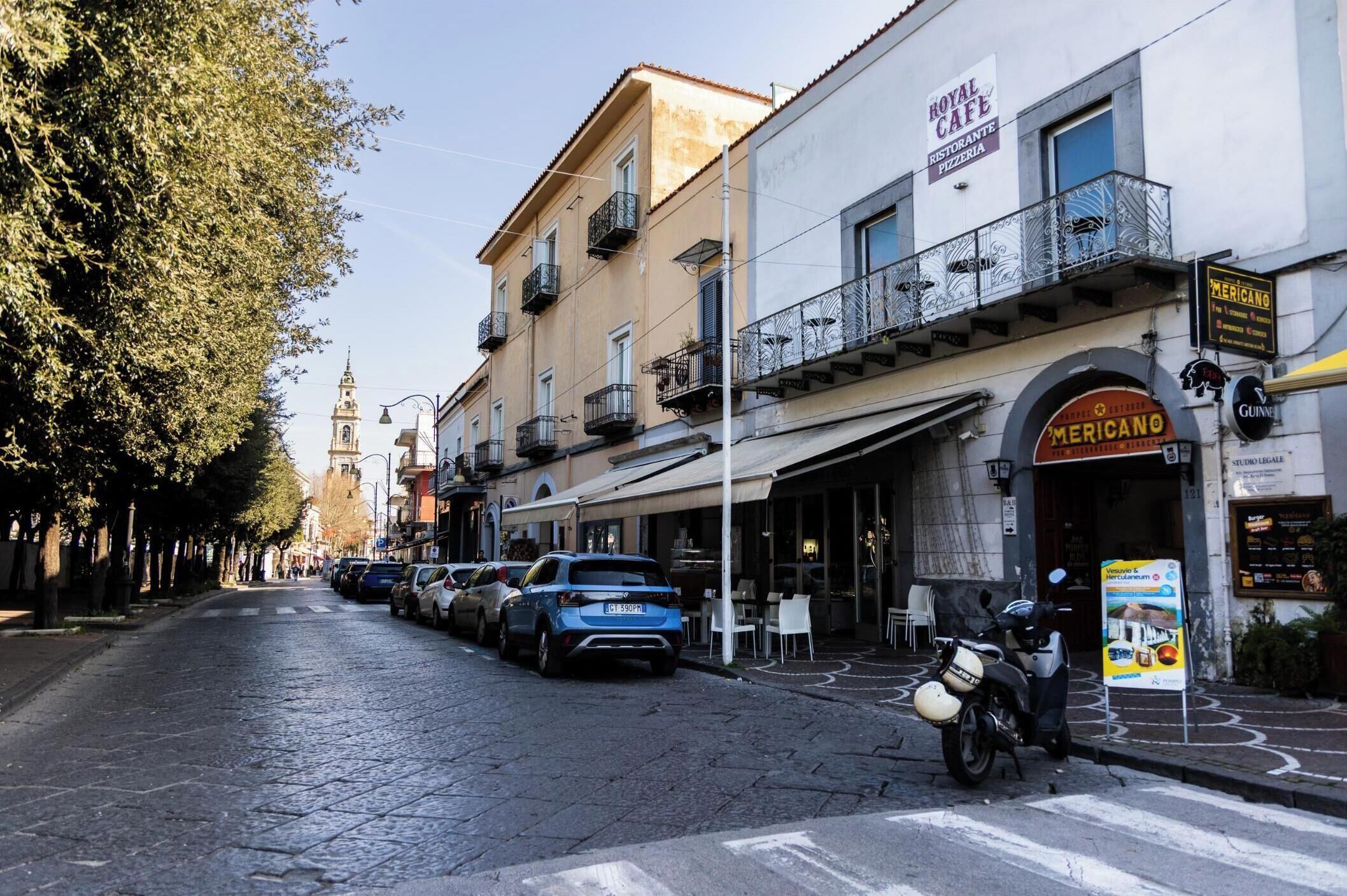 A street with parked cars and a motorcycle, buildings with balconies, and a sign for 'Mericano Restaurant'.