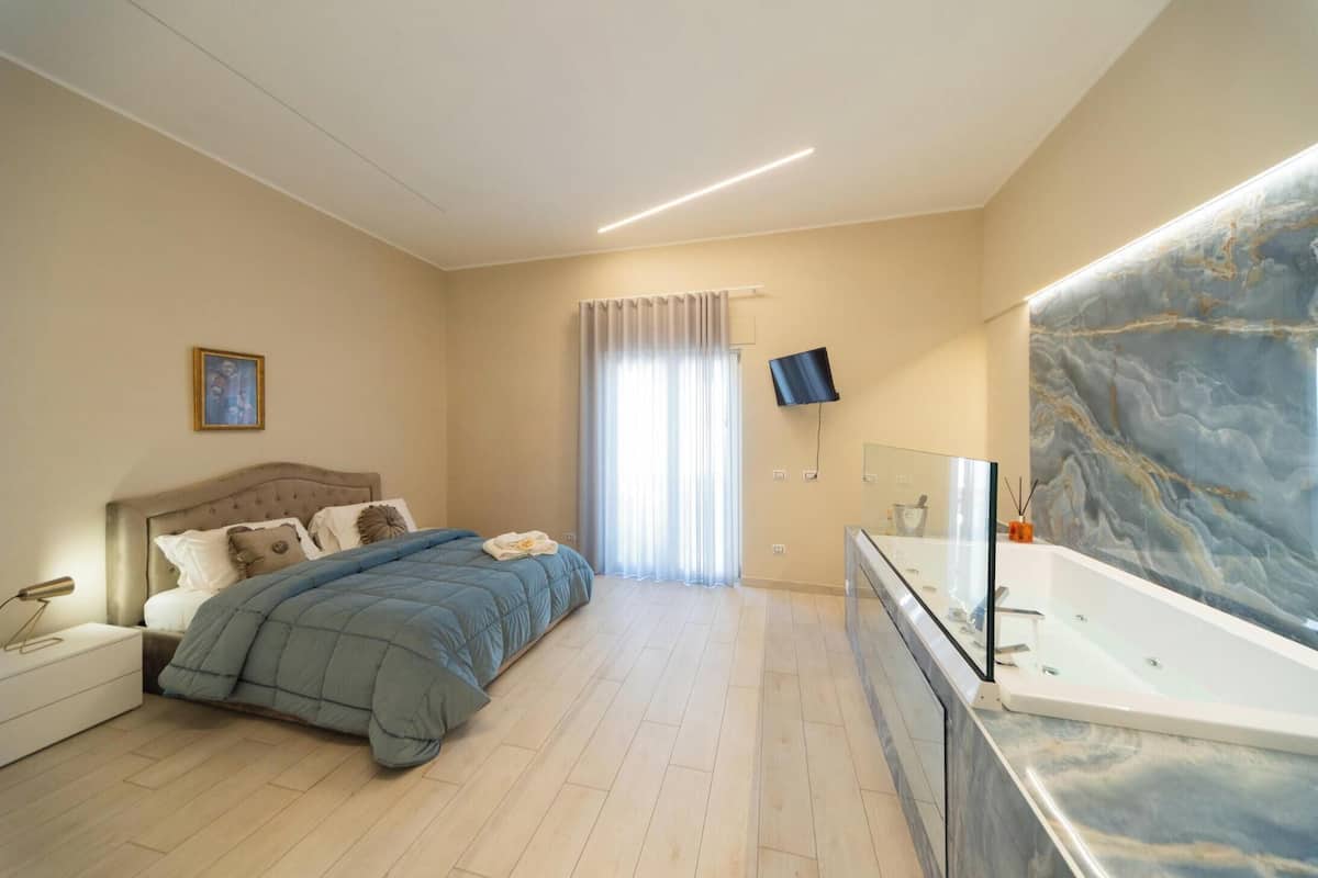 A bedroom with a bed, bedside table, TV, and a bathroom with a bathtub and mirror.