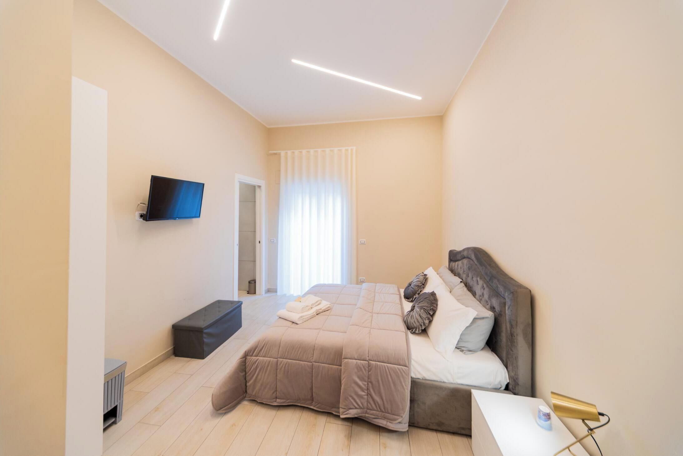 A modern bedroom with a large bed, a flat-screen TV mounted on the wall, and a window with sheer curtains.
