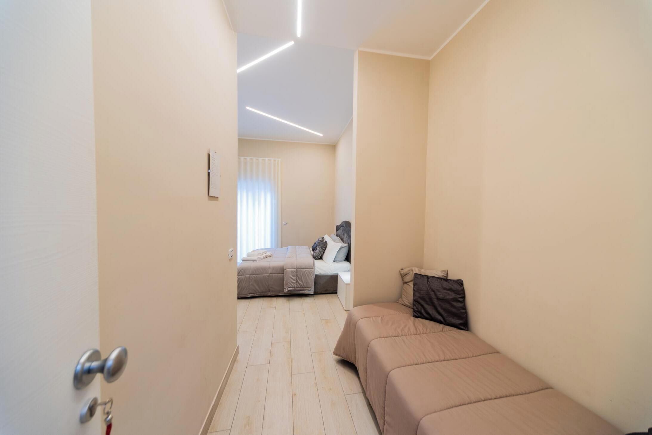 Una habitación de hotel estrecha con cama, un pequeño rincón de descanso y una ventana con persianas.