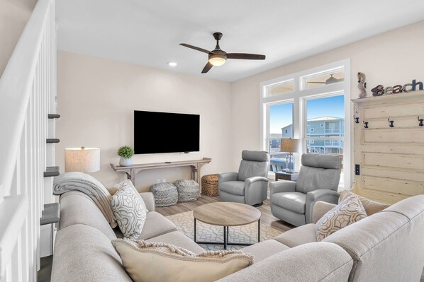 Ein modernes Wohnzimmer mit einem großen Flachbildfernseher, grauen Sofas, einem Couchtisch und einem Deckenventilator.