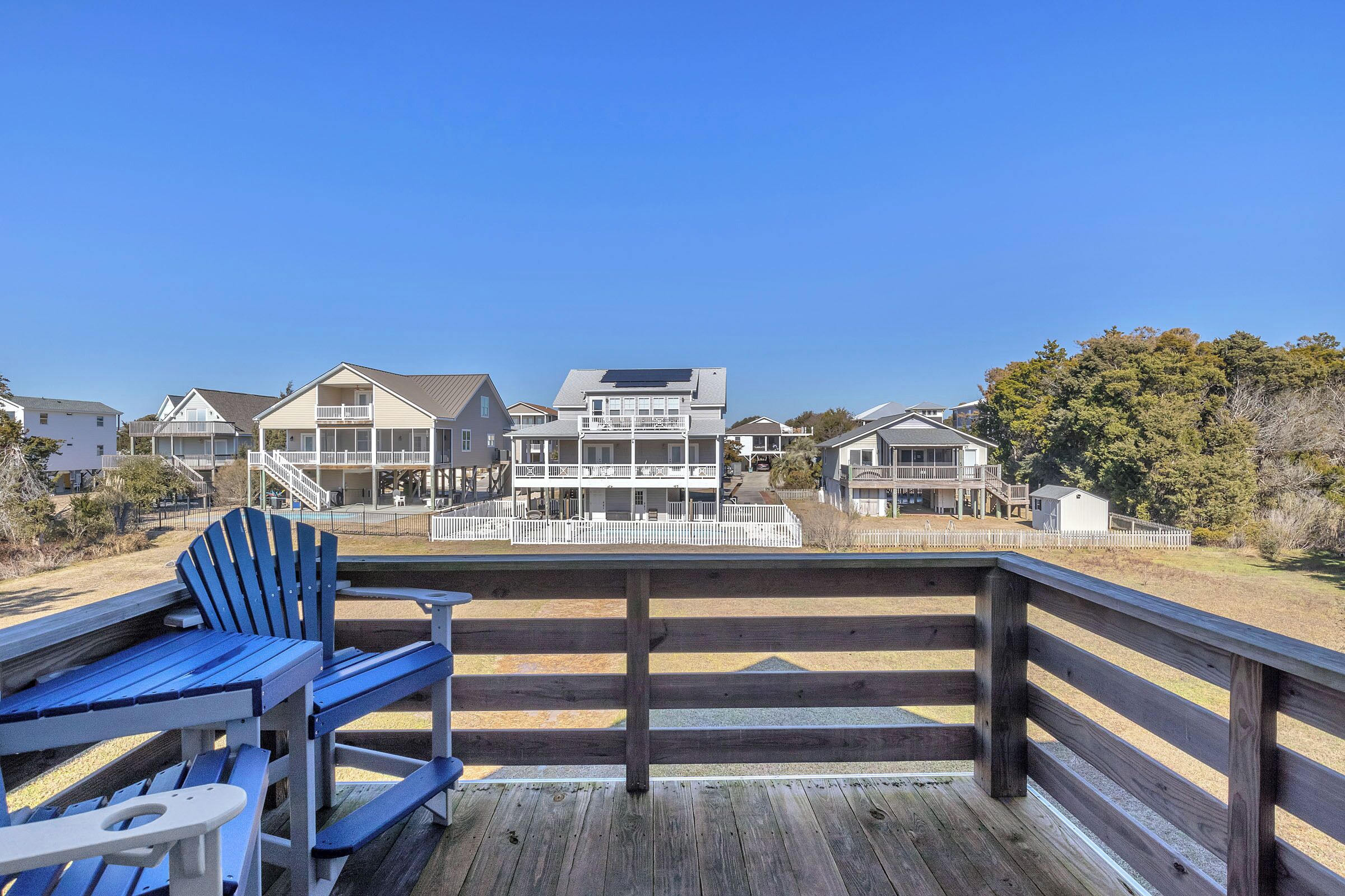 A wooden deck with a blue Adirondack chair and a white table, overlooking a residential area with houses and a clear blue sky.