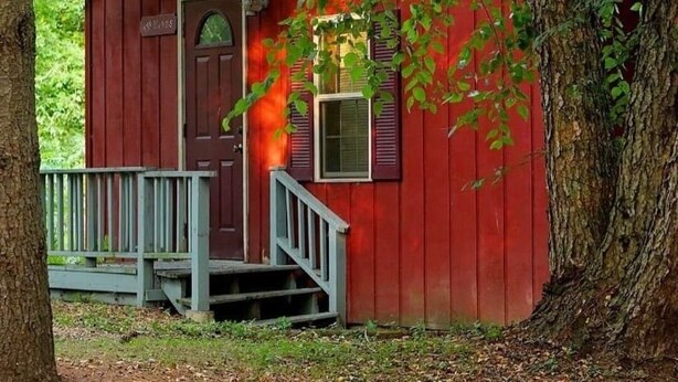 Une cabane rouge avec une porte en bois, une petite fenêtre et une véranda dotée d’une balustrade.