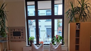 A neatly made bed with pillows and towels, a radiator, a window with blinds, a bookshelf, and two potted plants.