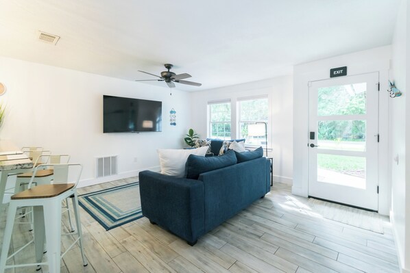 A modern living room with a blue sofa, a ceiling fan, a flat-screen TV, and a dining area with bar stools.