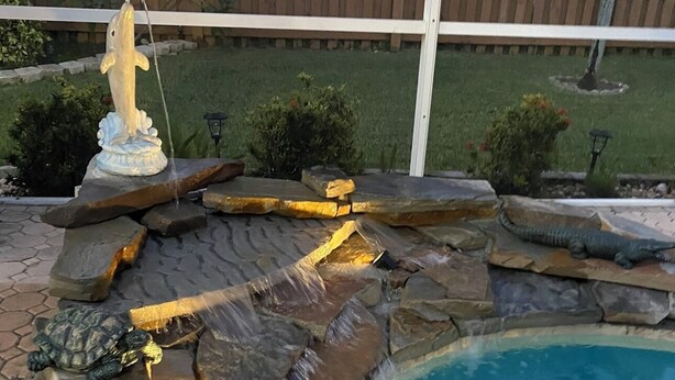 A swimming pool with a rock waterfall feature, a statue, and a wooden fence in the background. - Heated pool home. Desirable 3-bedroom located in CapeCoral loads of extras! (Cape Coral)
