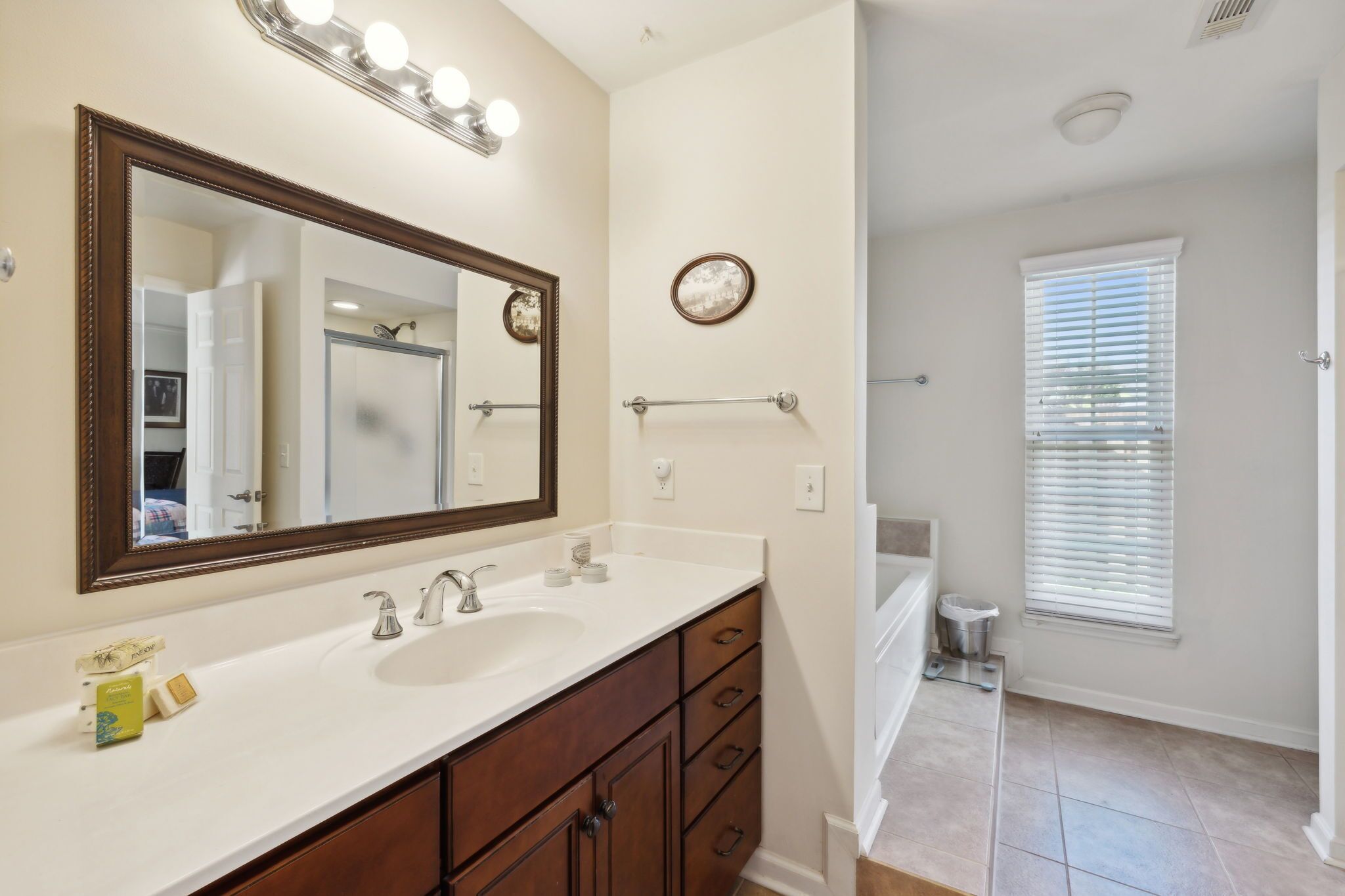 A bathroom with a large mirror, two sinks, a vanity with drawers, a towel rack, and a window with blinds.