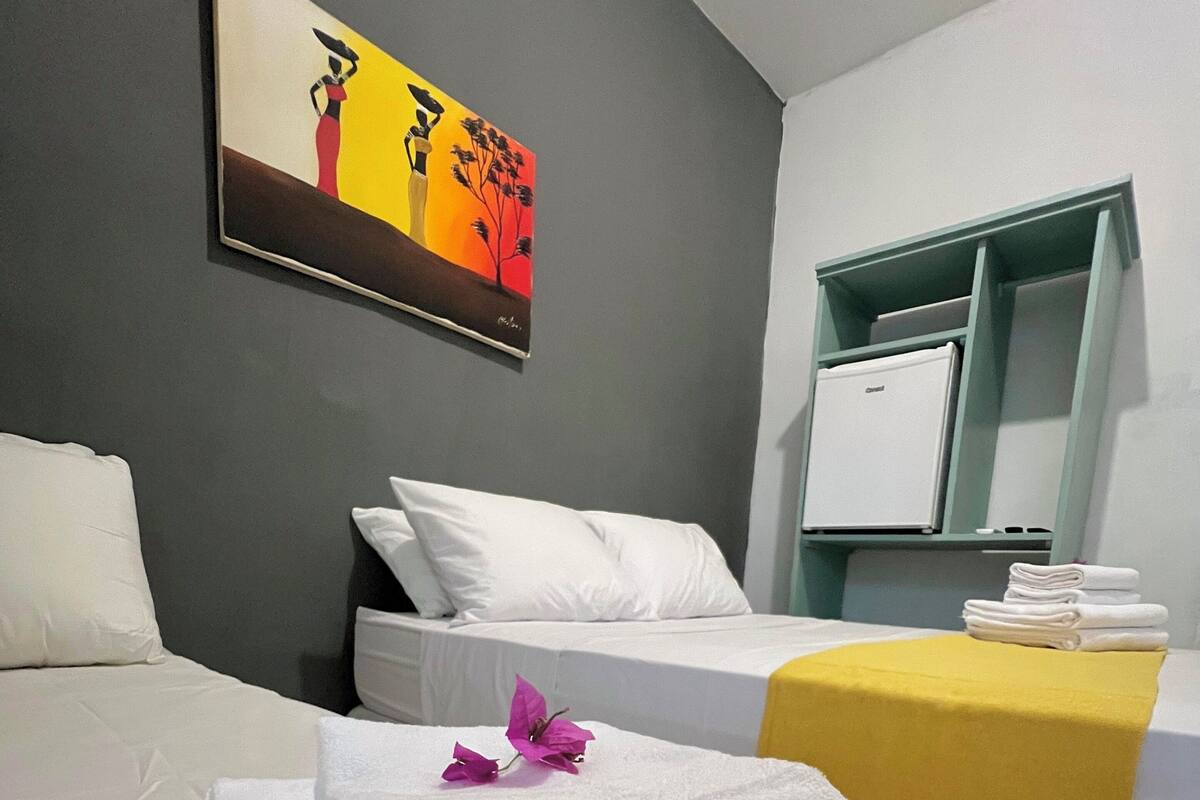 Habitación de hotel con dos camas, ropa blanca, una pared de acento amarilla, un cuadro y un minibar.