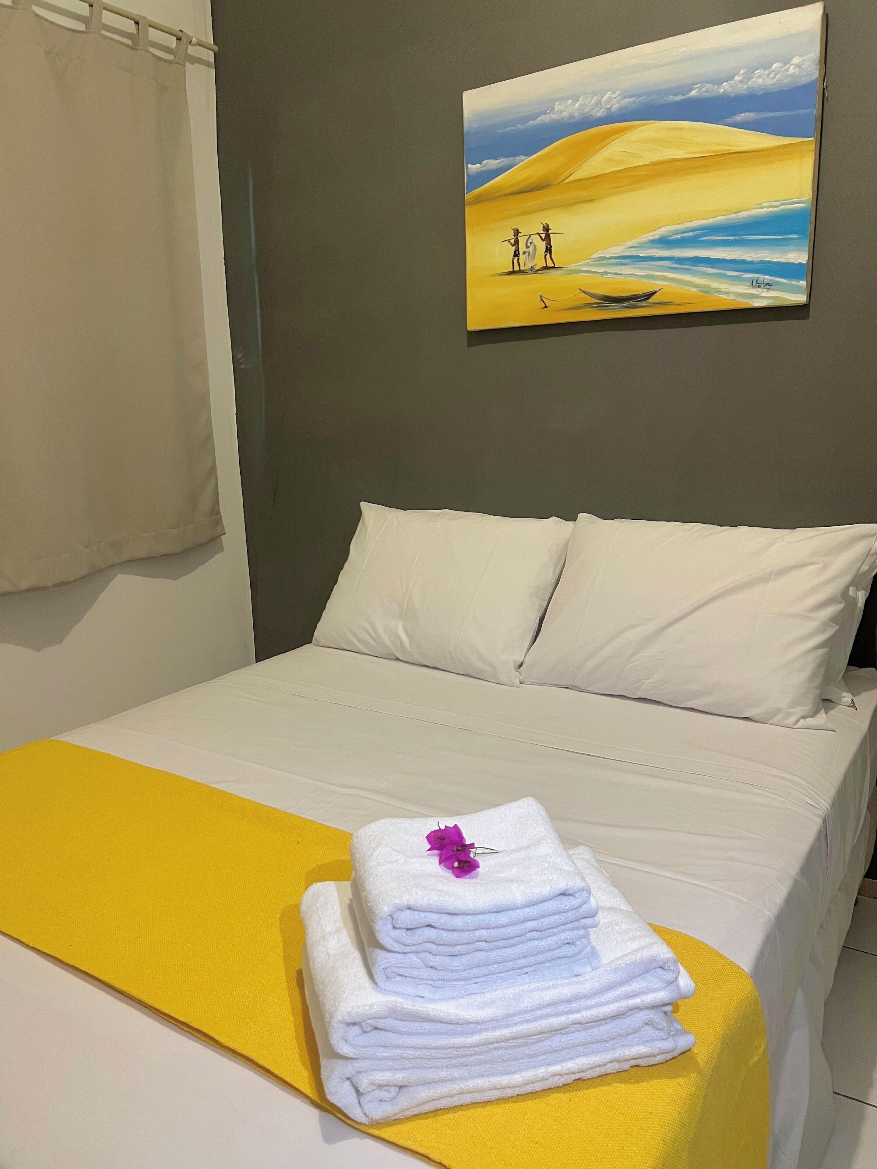 A neatly made bed with white linens and a yellow throw, a painting of a desert landscape on the wall, and a curtain on the window.