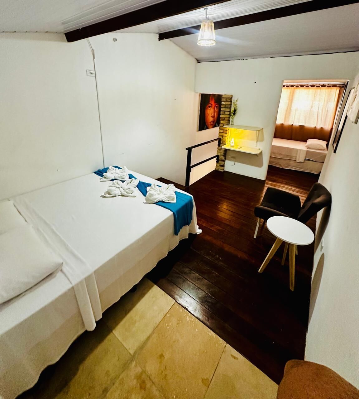 A small, compact room with a single bed, a small table, and a chair. There is a window with curtains, a wall-mounted shelf, and a light fixture.
