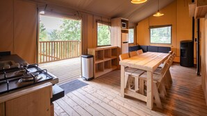 Un accogliente interno di una baita con un tavolo da pranzo in legno, panche, angolo cottura con fornello e un'ampia finestra che si affaccia sull'esterno.