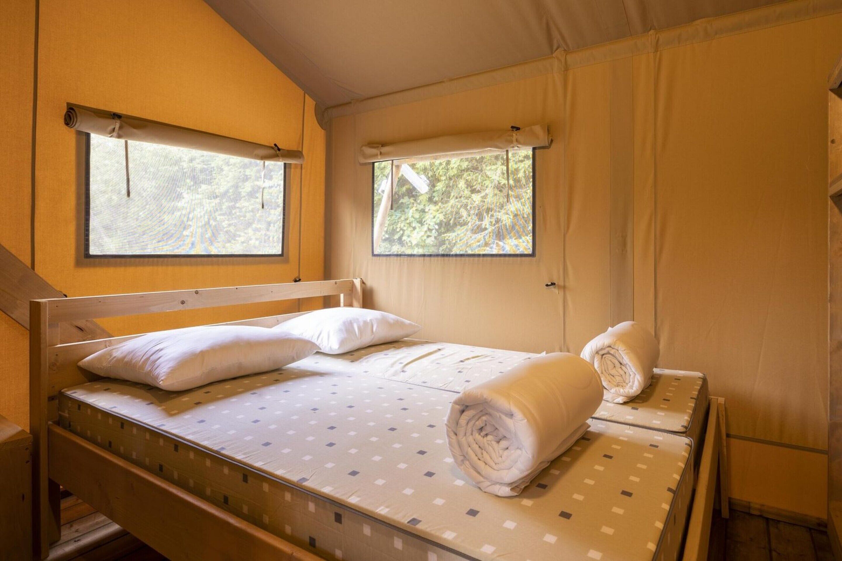 A single bed with a wooden frame, two pillows, and a rolled-up towel on a mattress with a patterned cover. There is a window with a view of greenery outside.