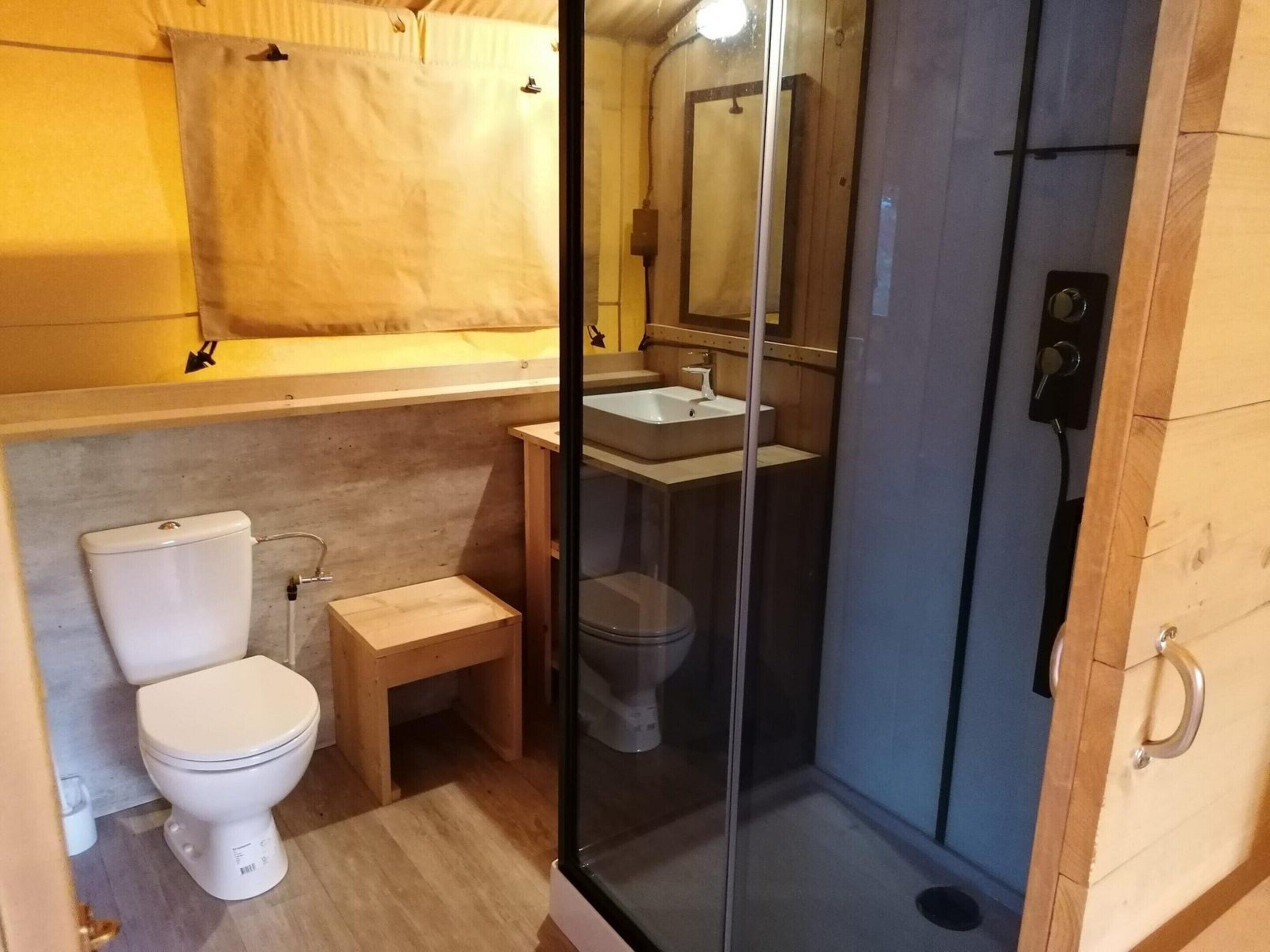 A compact bathroom with a toilet, a small wooden stool, a glass-enclosed shower, and a sink with a mirror above it.