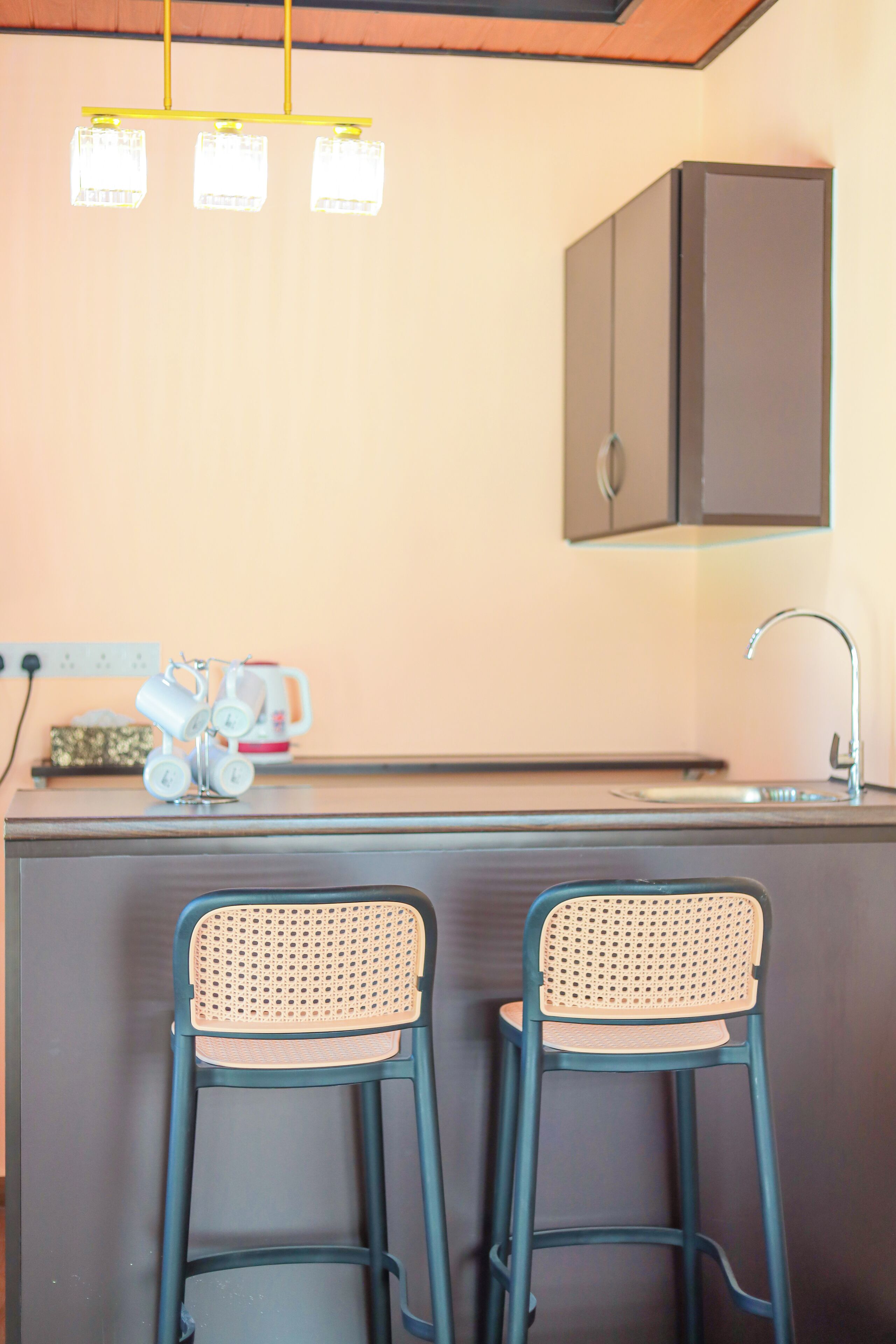 A kitchen with two bar stools, a cabinet, a coffee machine, and a hanging light fixture.