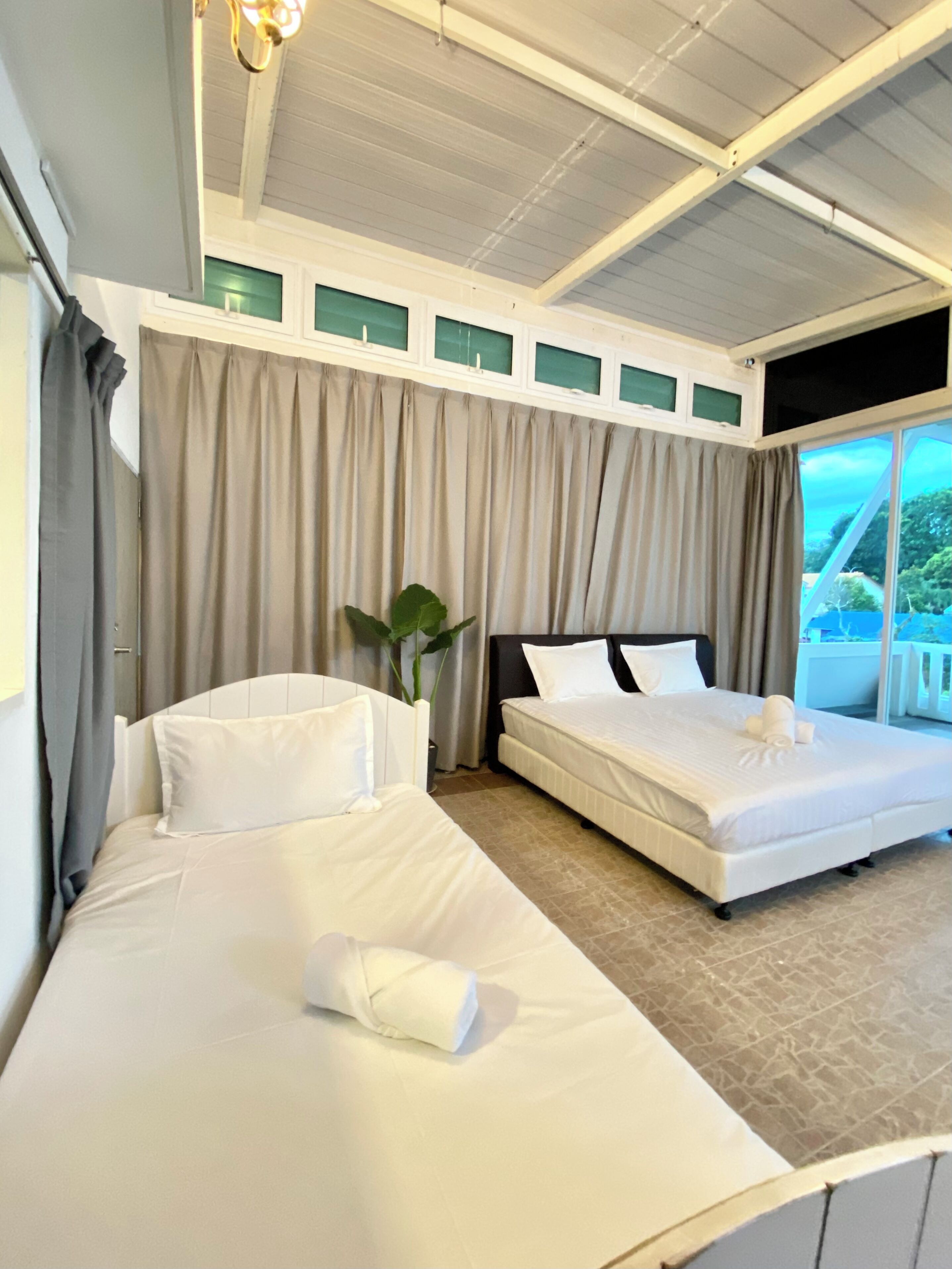 Quarto de hotel com duas camas, uma janela grande com cortinas, um ventilador de teto e um vaso de planta.
