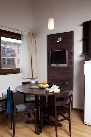 A compact living space with a round dining table, dark wooden chairs, a small TV mounted on a wooden panel, a white refrigerator, and a window with a view of buildings outside.
