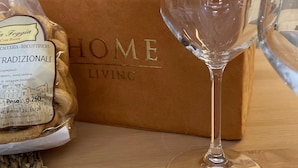 A glass of wine, a bag of biscotti, and a brown box labeled "HOME LIVING" on a wooden surface. - La mia Volta in Ostuni (Ostuni)