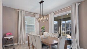 Een eetruimte met een houten tafel, zes stoelen, een hangende lamp en uitzicht buiten door grote ramen.