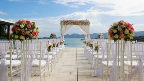 An outdoor wedding aisle with white chairs and floral arrangements, leading to a white arch with flowers, overlooking a body of water and mountains. - Samujana Villas - Villa Karawek (Bo Put)