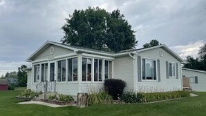 A single-story mobile home with a covered porch, a small front yard, and a large tree in the background.