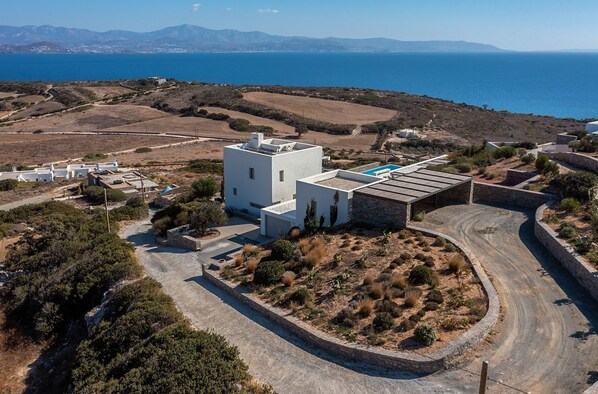A modern white building with a pool, surrounded by dry vegetation and a winding road, overlooking a coastal landscape.