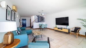 A modern living room with a sofa set, a dining table, a TV, and a staircase leading to an upper level.
